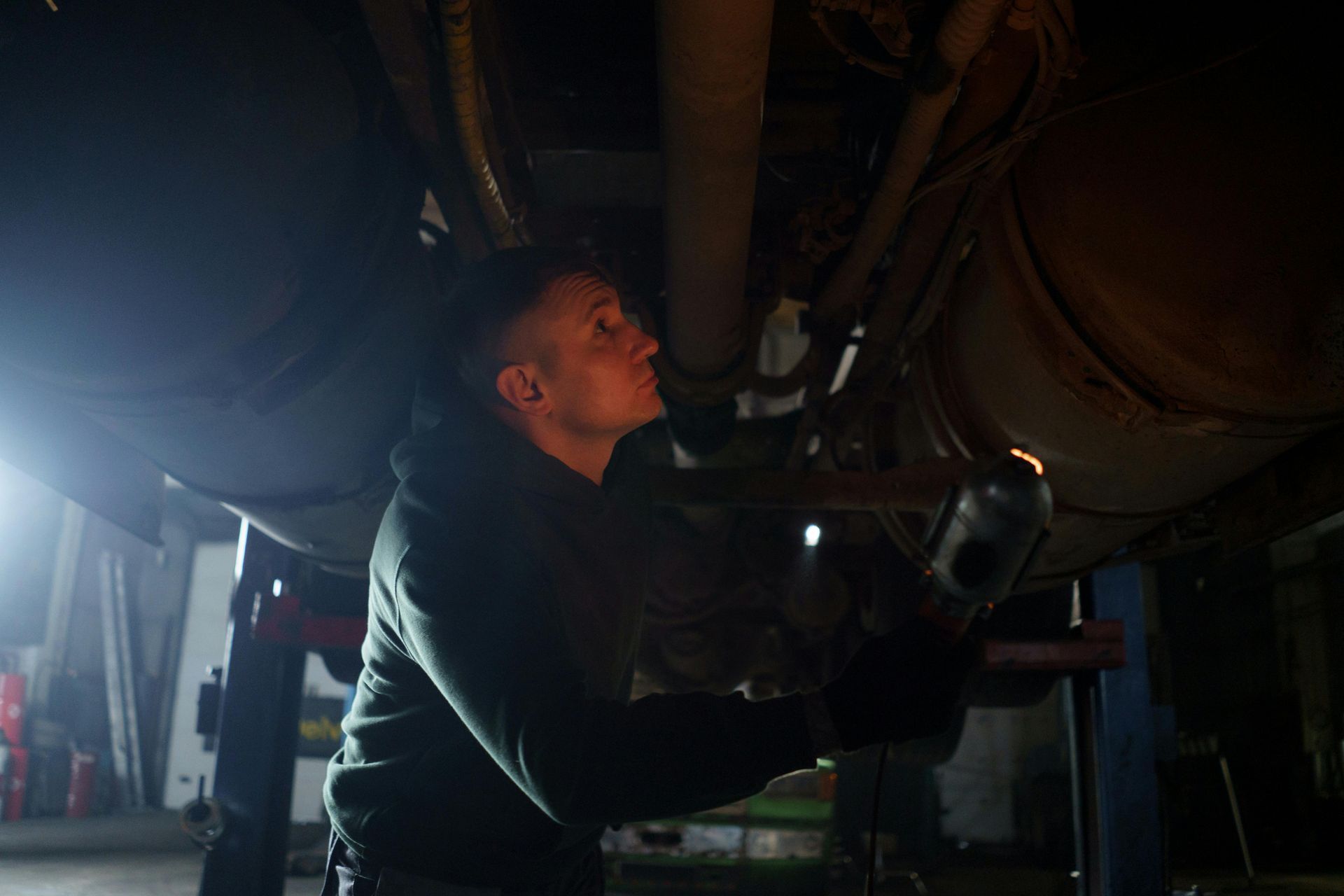 Mechanic works under a vehicle, inspecting parts, illuminated by a work light, in a garage.
