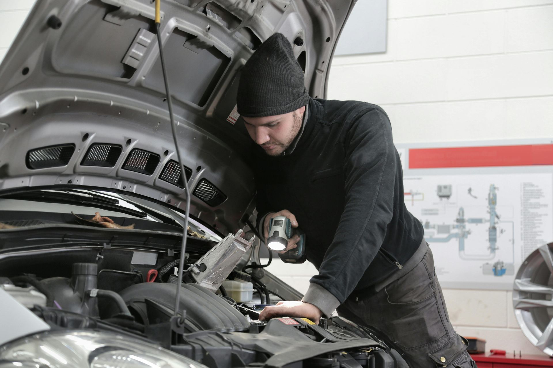 Mechanic inspecting a car engine, holding a flashlight. The mechanic wears a black beanie and is in a garage.