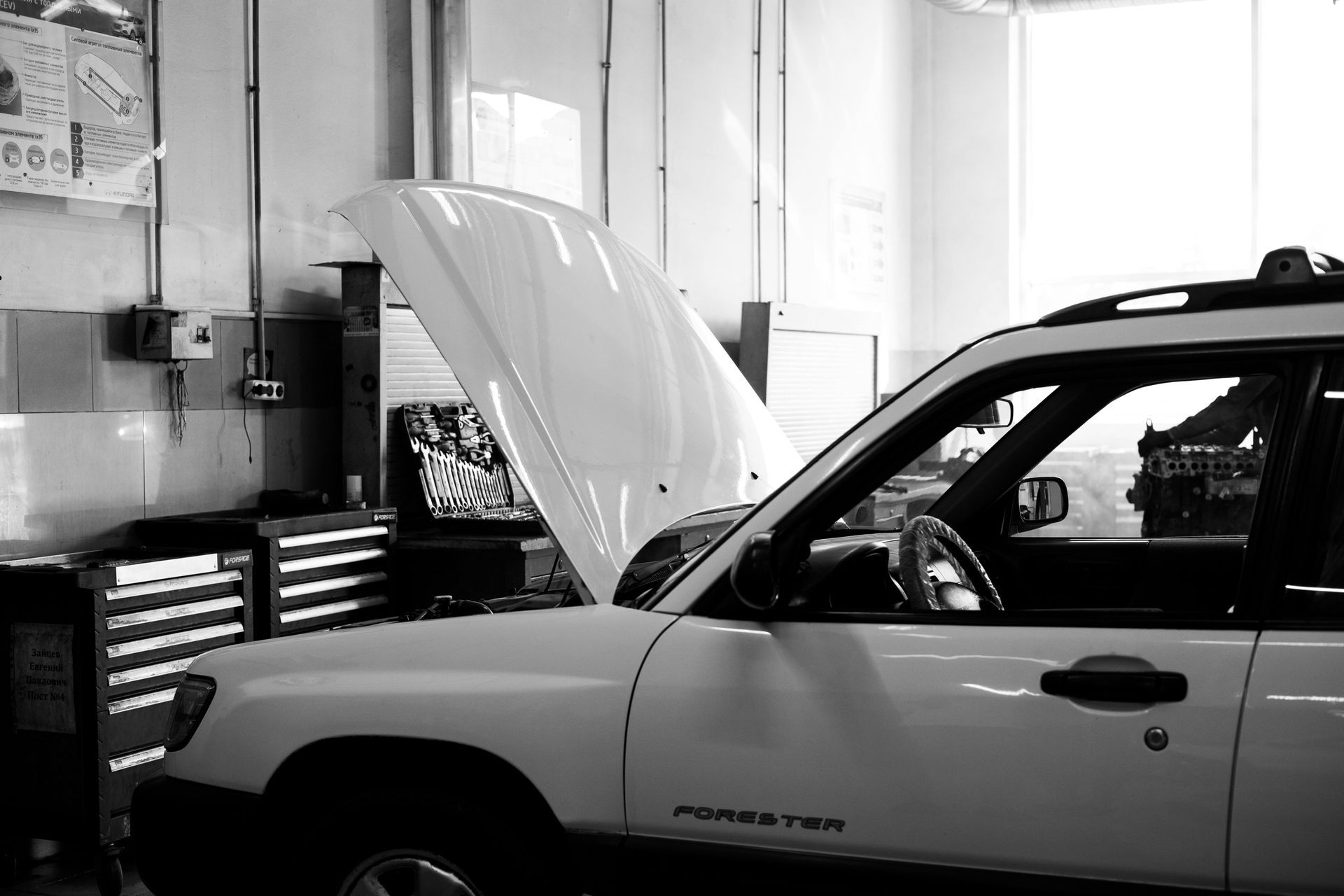 White car with hood up in auto repair shop; tools and cabinets visible.