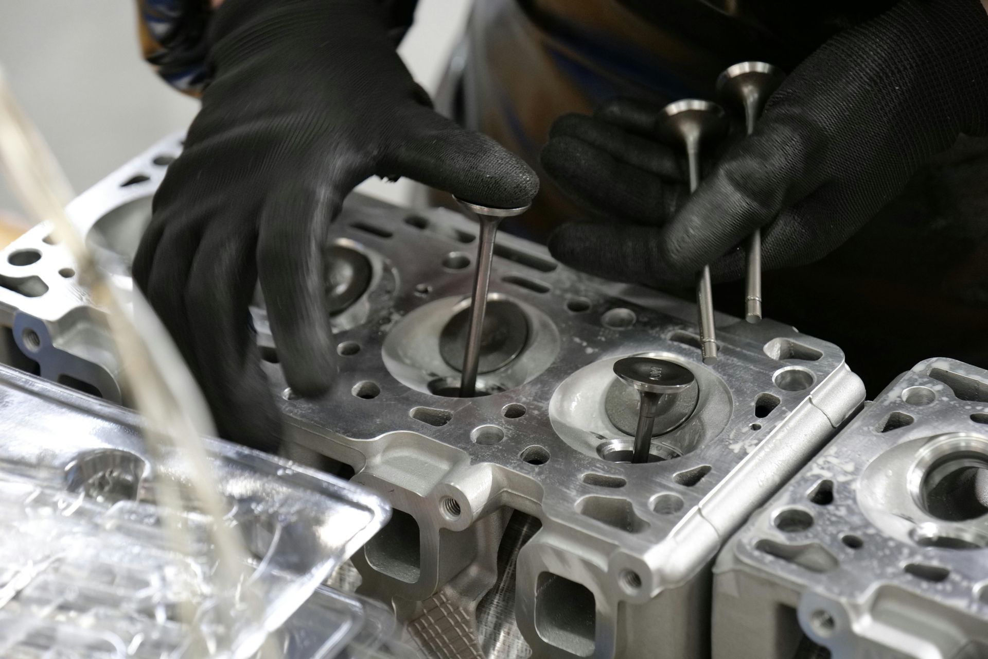 Gloved hands installing valve stems into a metal cylinder head, likely for an engine, at a manufacturing facility.
