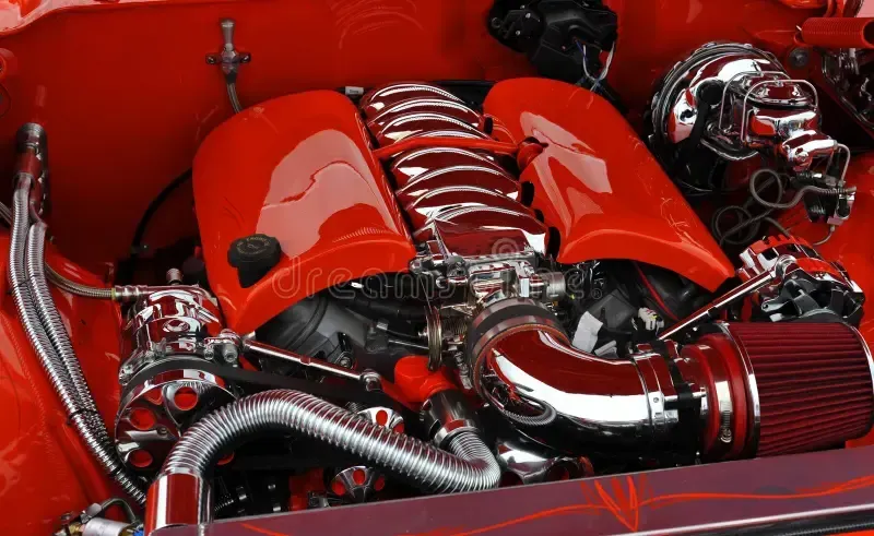 Red and chrome engine in a car, with intake and headers.