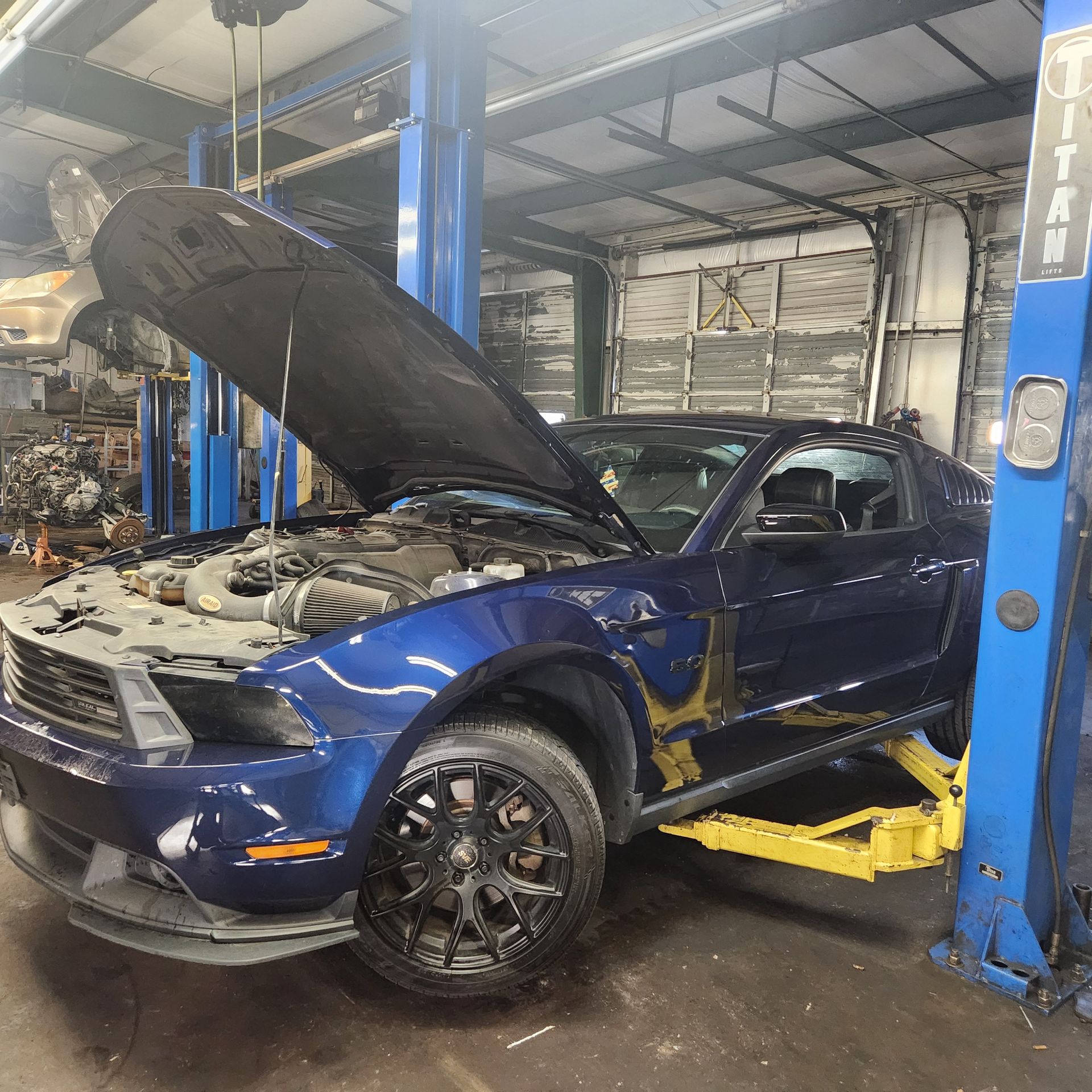 Blue Ford Mustang on a lift in a repair shop with its hood open for service.
