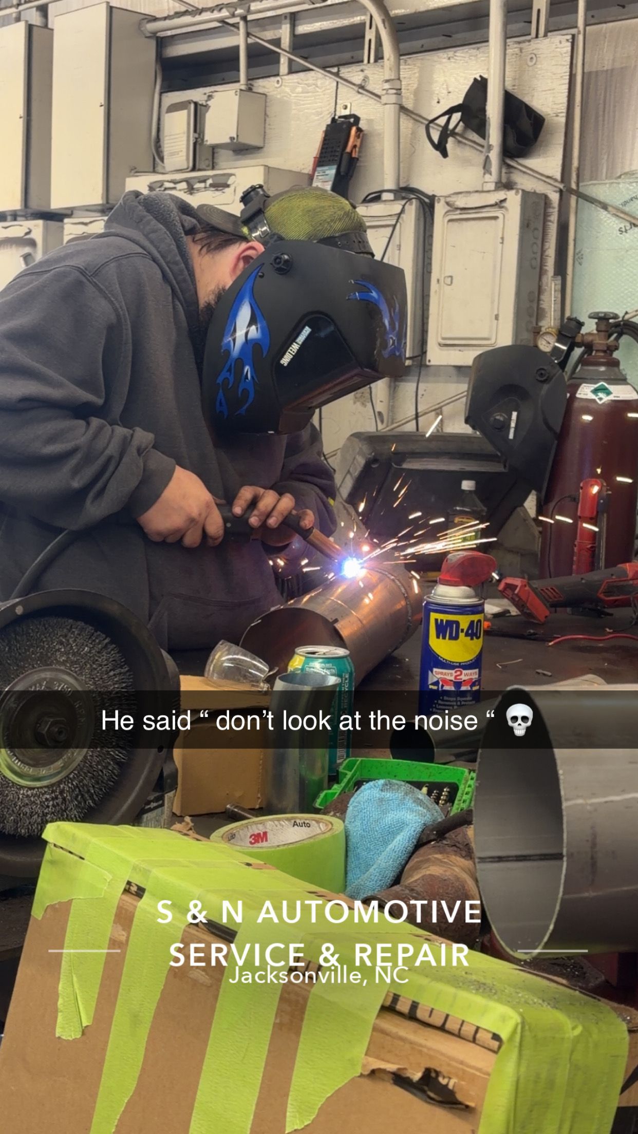 Person welding in a garage; sparks fly. A blue welding helmet is worn. A sign for 