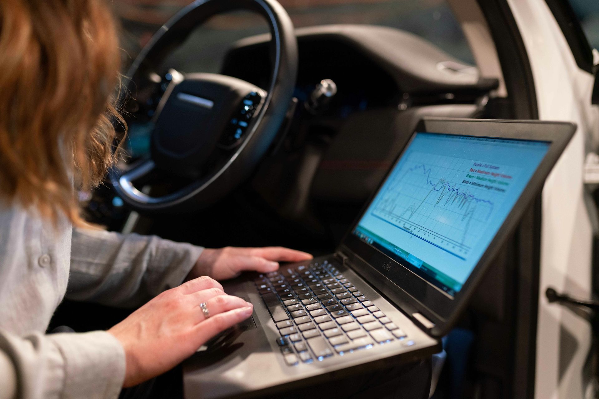Person using laptop inside a car, likely for diagnostics. Screen shows data. Steering wheel visible.